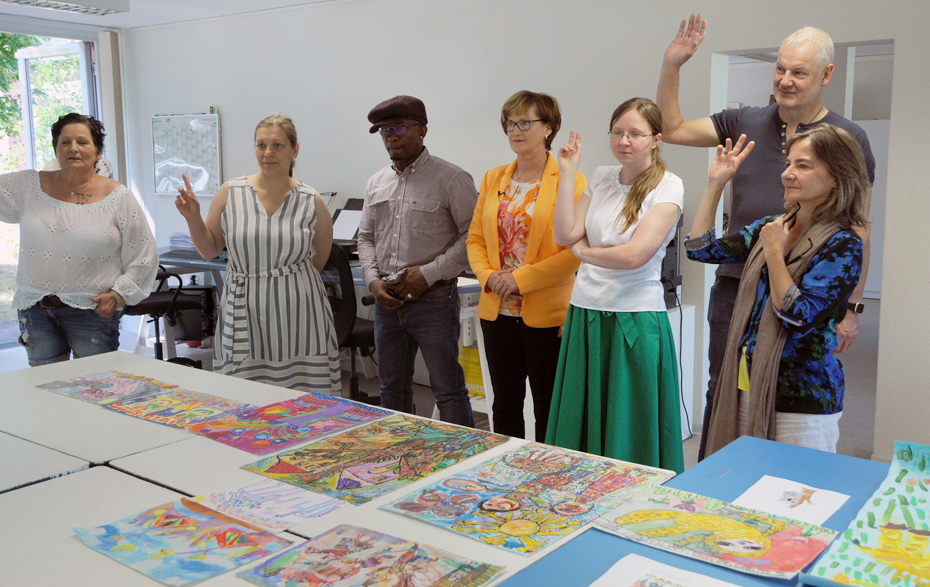 Foto: 7 Personen stehen an einem Tisch mit Zeichenarbeiten und heben teilweise die Hand Foto: 7 Personen stehen an einem Tisch mit Zeichenarbeiten und heben teilweise die Hand