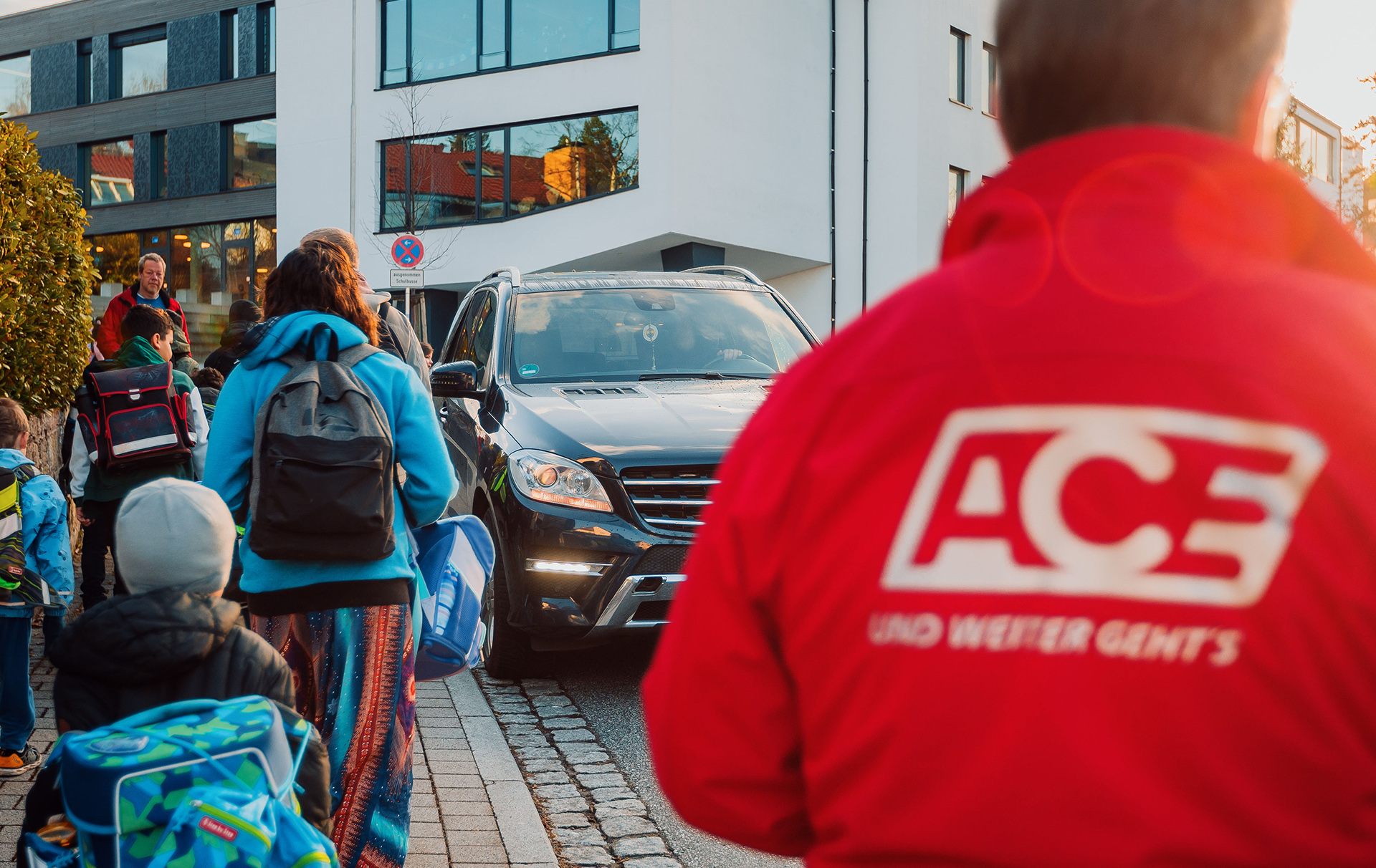 Foto: Schulkinder laufen an einem Auto vorbei, ein Man in roter Jacke beobachtet das Foto: Schulkinder laufen an einem Auto vorbei, ein Man in roter Jacke beobachtet das