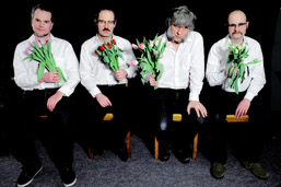 Foto: Die 4 Bandmitglieder sitzen mit weißen Hemden, schwarzen Hosen und jeweils einem Tulpenstrauß in einer Hand auf Holzstühlen in einer Reihe. Foto: Die 4 Bandmitglieder sitzen mit weißen Hemden, schwarzen Hosen und jeweils einem Tulpenstrauß in einer Hand auf Holzstühlen in einer Reihe.