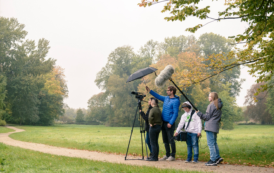 Foto: Gruppe beim Filmen mit Stativ, Kamera, Mikrofon, Aufnahmetasche und Schirm n einer Parklandschaft