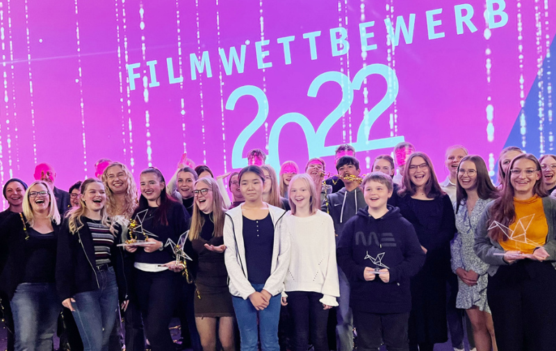 Gruppenfoto vor pinker Leinwand mit dem Text „Filmwettbewerb 2022“ Gruppenfoto vor pinker Leinwand mit dem Text „Filmwettbewerb 2022“